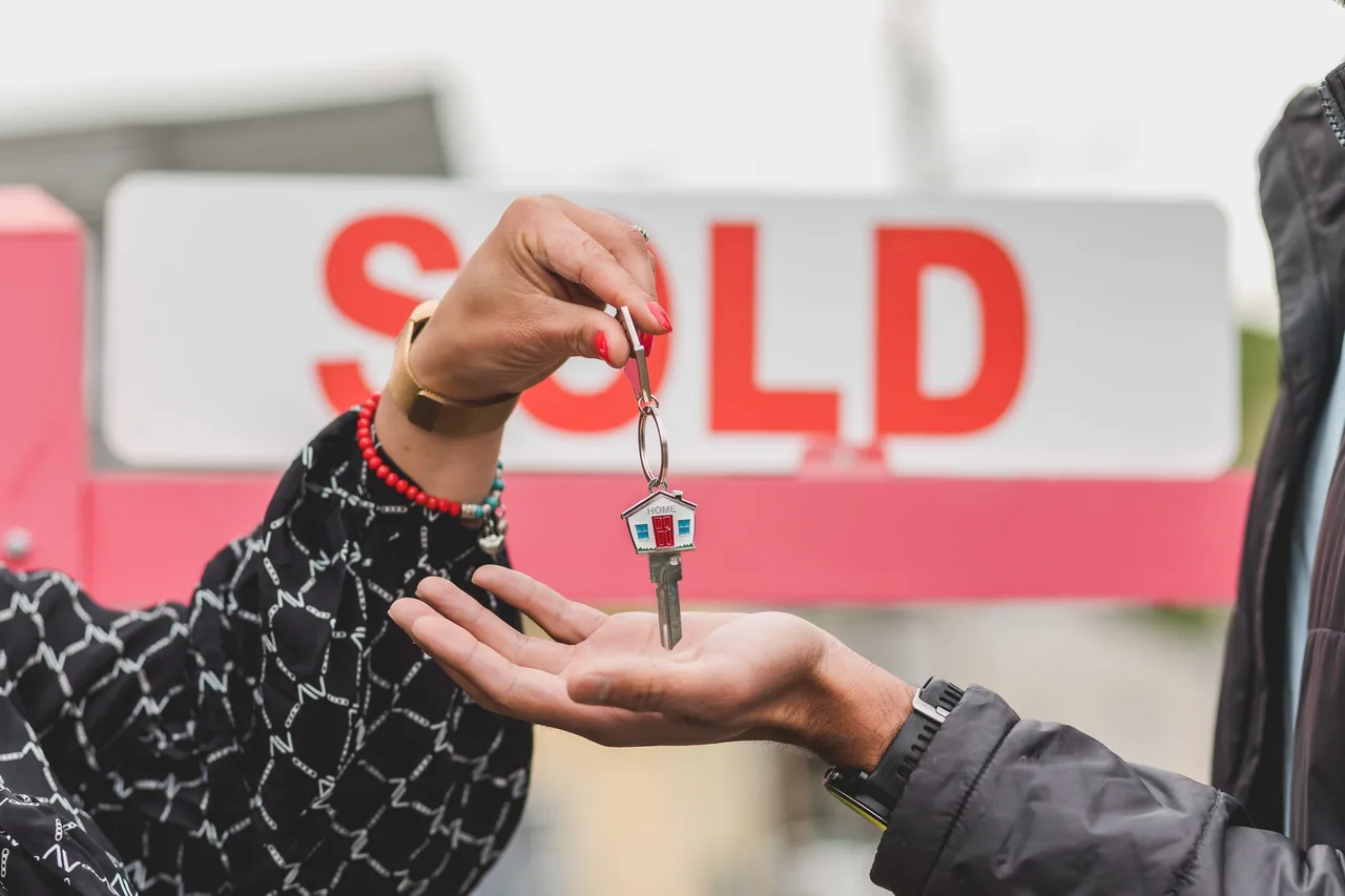 Close-up of handing over house key with sold sign in background, symbolizing real estate success