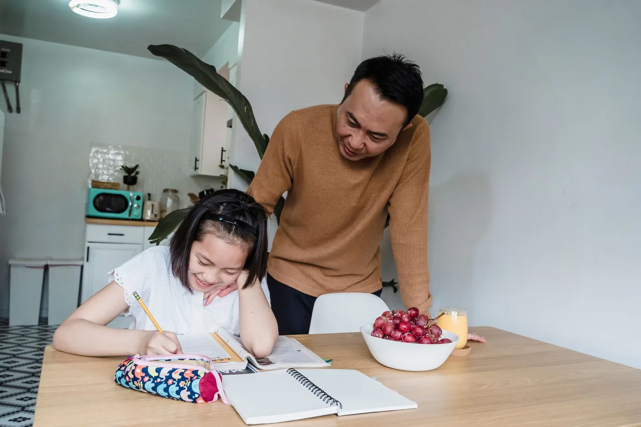 Asian father helping his daughter with homework at the kitchen table, fostering education and smiles