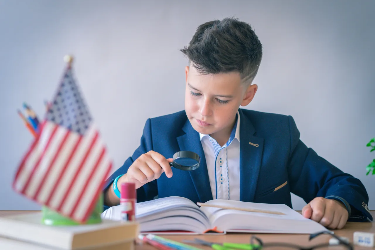 Young boy in a blue suit studying with a magnifying glass next to a desk with the US flag indoors