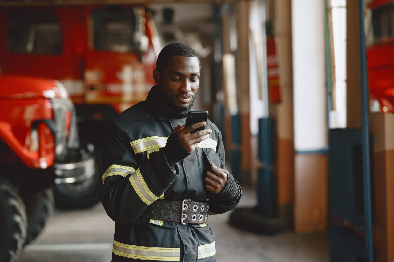 Firefighter in uniform checking his smartphone inside a fire station with fire trucks in the background