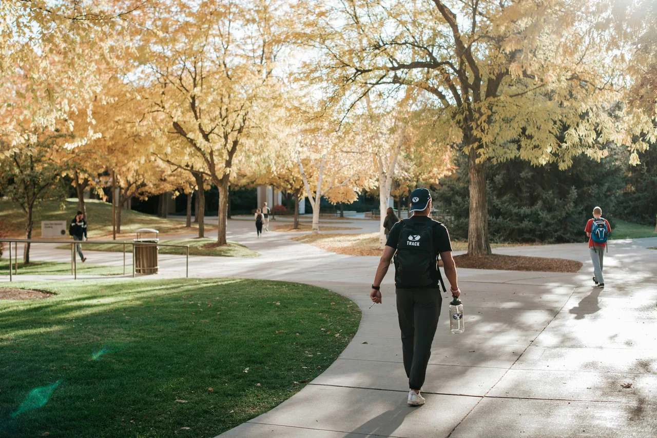A man with a track backpack walks across the BYU campus in autumn