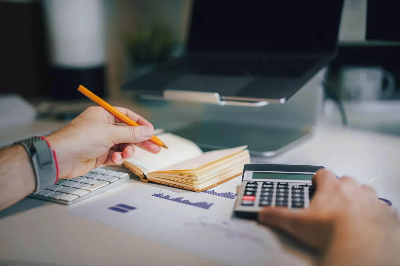 Hands using calculator and taking notes on desk with laptop, showcasing business analytics