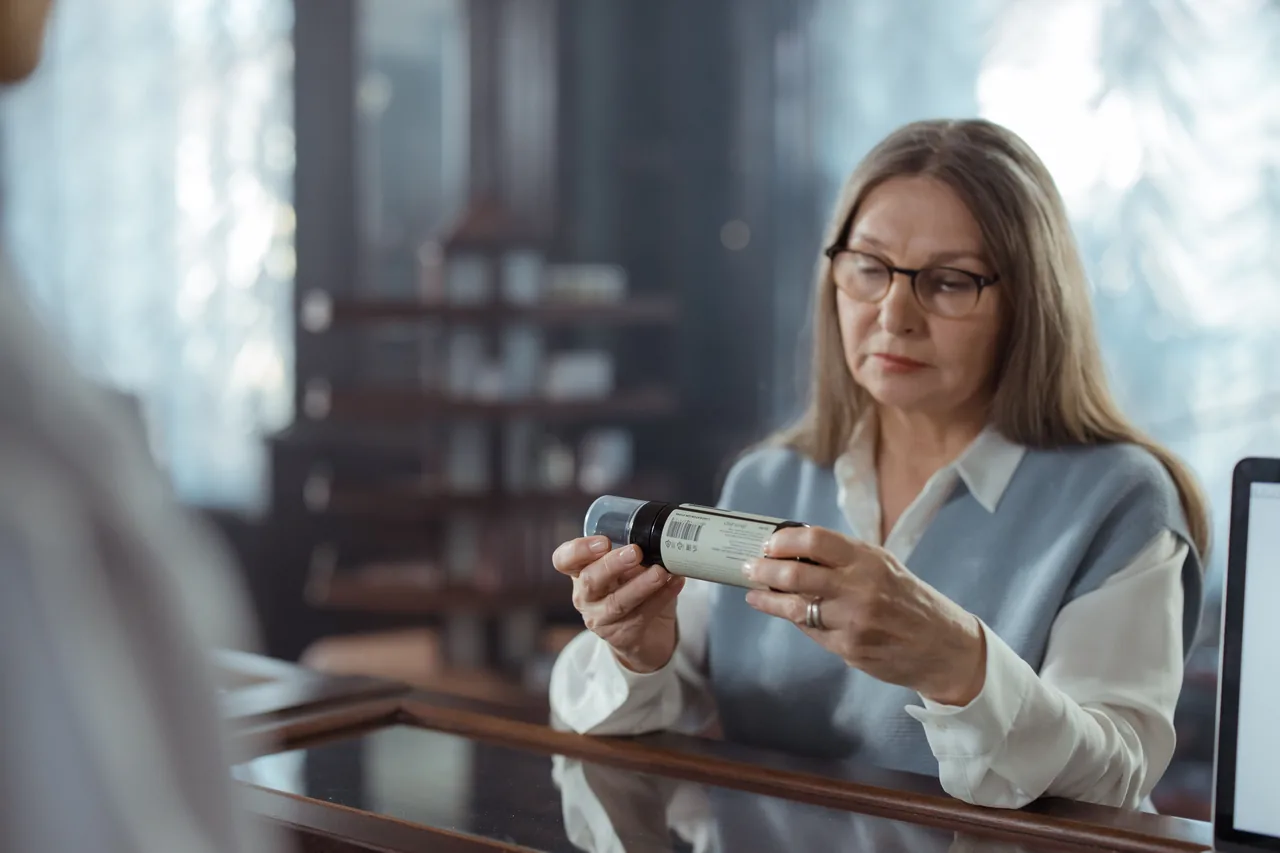 A senior woman with eyeglasses reviews a medicine bottle at a pharmacy counter indoors