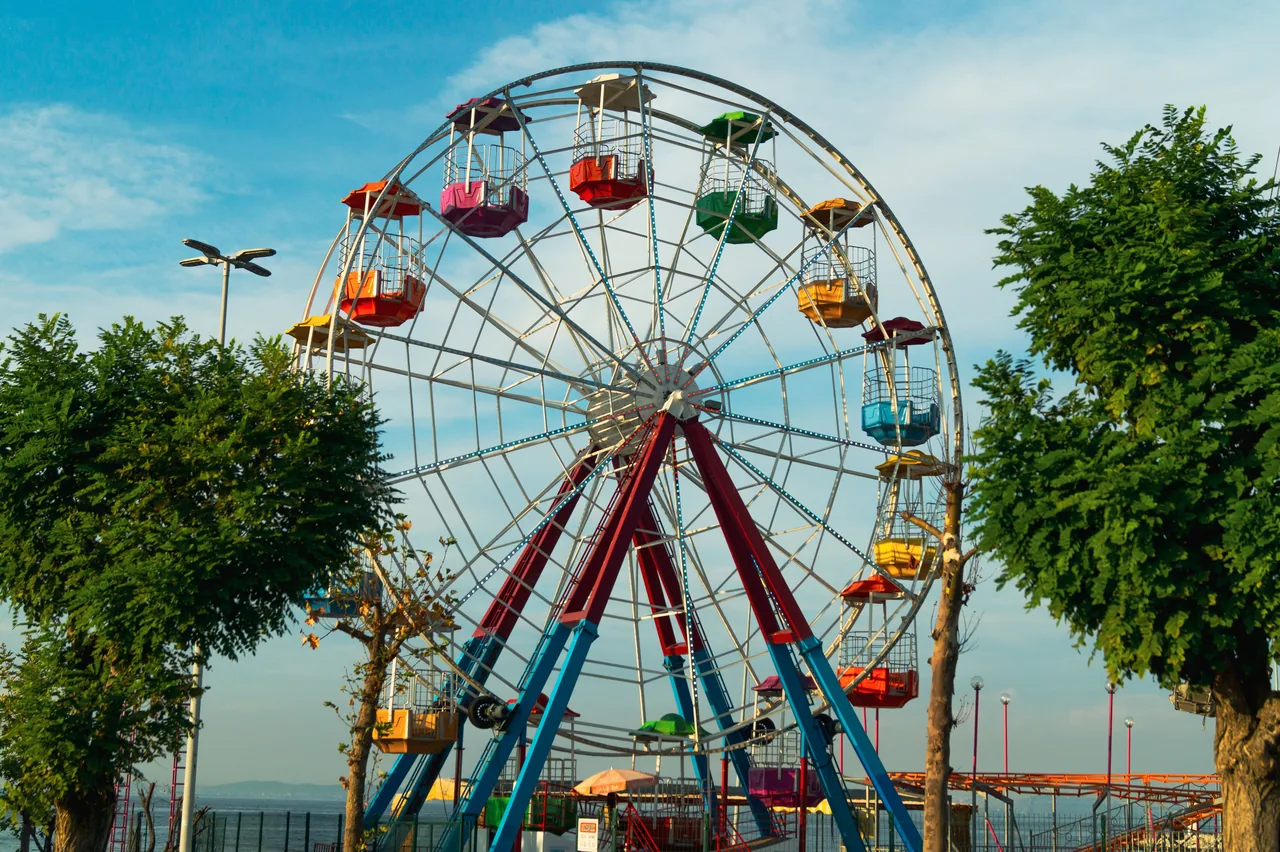 Colorful Ferris wheel at an outdoor amusement park during daytime, surrounded by trees and blue sky